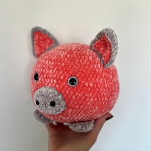 May include: A plush, round pig toy in shades of coral and gray. The pig has a textured, velvety appearance, with gray accents on the ears, snout, and feet. The eyes are black with silver glitter. A handmade, soft toy.
