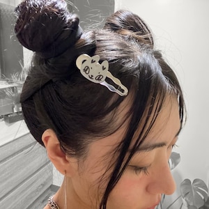 Puede incluir: Una pinza para el pelo plateada con un dise&ntilde;o de cara de gato. La cara del gato tiene dos corazones como ojos y el texto "Dear" escrito en ella.