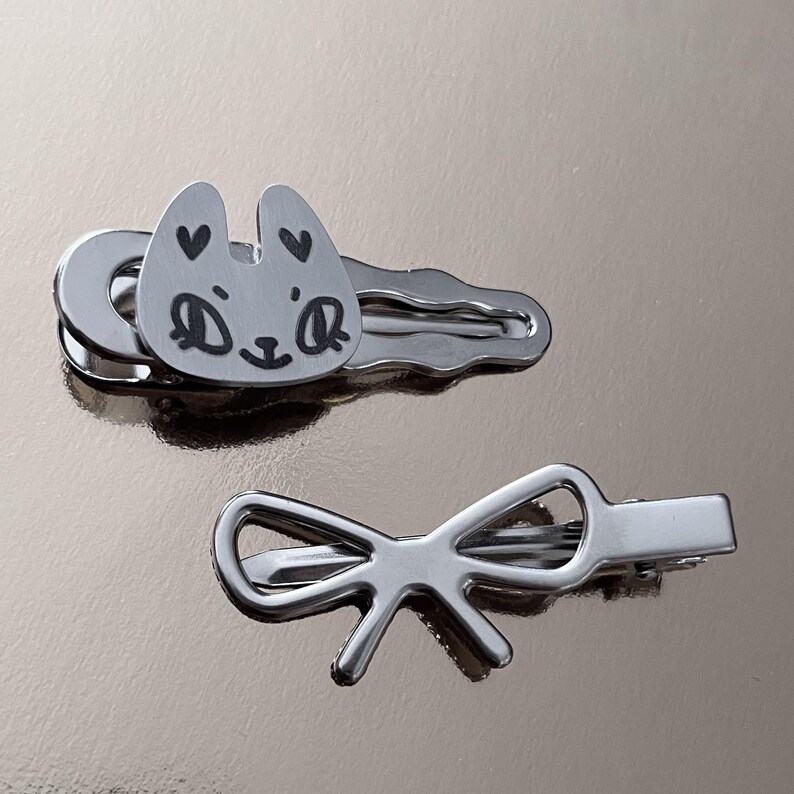 Puede incluir: Dos pinzas para el pelo plateadas. Una pinza tiene un rostro de gato de dibujos animados con corazones como orejas. La otra pinza tiene forma de lazo.