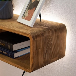 May include: A wooden wall shelf with a curved front edge and a built-in shelf. The shelf is lit with a strip of LED lights that can be controlled with a remote. The remote has buttons for on/off, brightness, and flashing modes. The shelf is mounted on a white wall and has a picture frame and a potted plant on top.