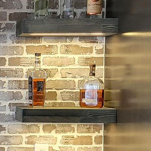 Può includere: Un allestimento di bar domestico con due mensole in legno scuro contro un muro di mattoni. La mensola superiore contiene bottiglie di liquore, tra cui Tito's Handmade Vodka e Svedka Vodka. La mensola inferiore mostra due bottiglie di liquore color ambra.