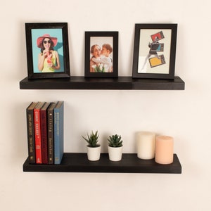 May include: Two black floating shelves with a variety of items on them, including framed photos, books, candles, and small plants. The shelves are mounted on a white wall.