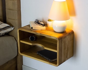 May include: A wooden wall-mounted nightstand with two shelves. The top shelf has a book, glasses, and a small potted plant. The bottom shelf has a watch and a book. A lamp with a white shade is on the top shelf.
