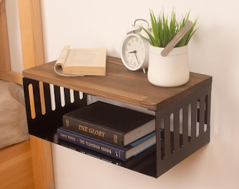 May include: A black metal floating shelf with a wood top. The shelf is mounted to a white wall and has a book, a clock, and a potted plant on it. The shelf is approximately 38 cm wide and 15 cm deep.