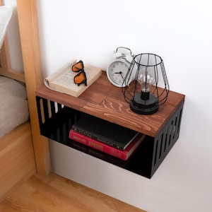 May include: A black floating shelf with a wooden top, featuring a book, sunglasses, a white alarm clock, and a black caged lamp. The shelf is mounted on a white wall next to a wooden bed frame.