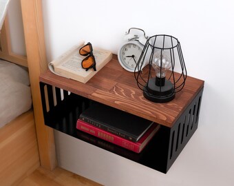 May include: A black floating shelf with a wooden top, featuring a book, sunglasses, a white alarm clock, and a black caged lamp. The shelf is mounted on a white wall next to a wooden bed frame.