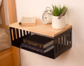 May include: A black metal wall shelf with a wooden top. The shelf has a slatted design and is holding a book, a clock, and a potted plant. The shelf is mounted on a white wall.