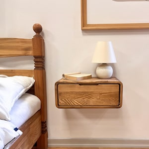 May include: A floating wooden nightstand with a single drawer and rounded edges. A small white lamp with a beige lampshade and a book rest on top. The nightstand is mounted on a light-colored wall next to a wooden bed frame.