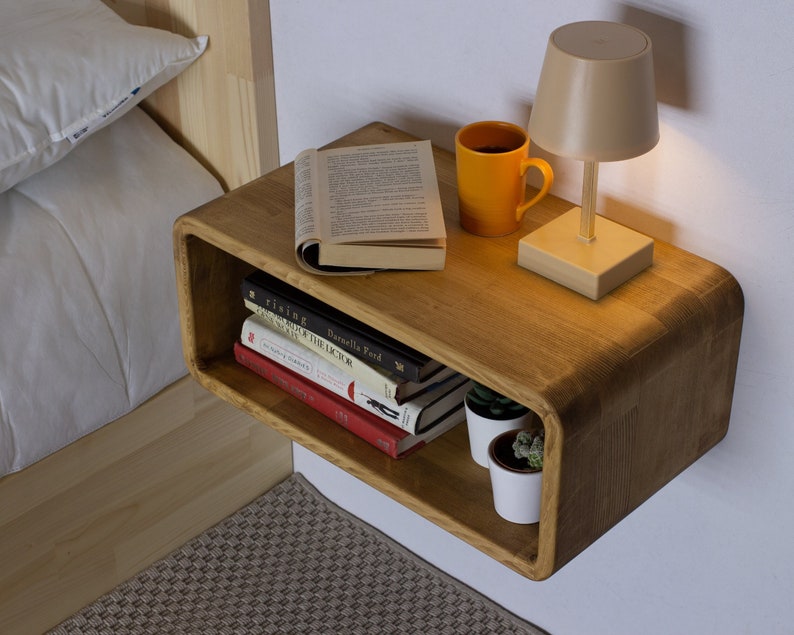 May include: A wooden wall-mounted nightstand with a curved edge. The nightstand has a shelf and a top surface. There are books, a lamp, a mug, and two small potted plants on the nightstand.