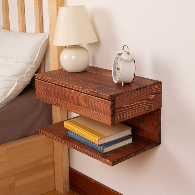 May include: A wall-mounted floating nightstand crafted from dark stained wood with a visible grain pattern. The unit features a top drawer and an open lower shelf. A white table lamp and a white analog alarm clock sit on the top surface.
