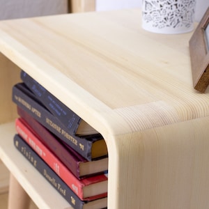 May include: A light wood nightstand with rounded edges and a shelf. The shelf is holding a stack of books with titles visible, including "The Writing After" and "Suzanne Forster 71".