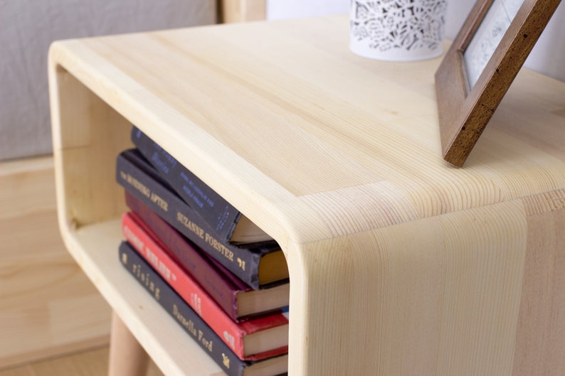 May include: A light wood nightstand with rounded edges and a shelf. The shelf is holding a stack of books with titles visible, including "The Writing After" and "Suzanne Forster 71".