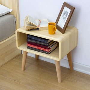 May include: A light wood nightstand with a curved front and rounded edges. It has a shelf for books and a small open space for a cup or other items. The nightstand has four legs and is next to a bed with a white pillow and a gray blanket.