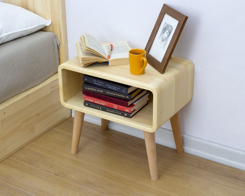 May include: A light wood nightstand with a curved front and rounded edges. It has a shelf for books and a small open space for a cup or other items. The nightstand has four legs and is next to a bed with a white pillow and a gray blanket.