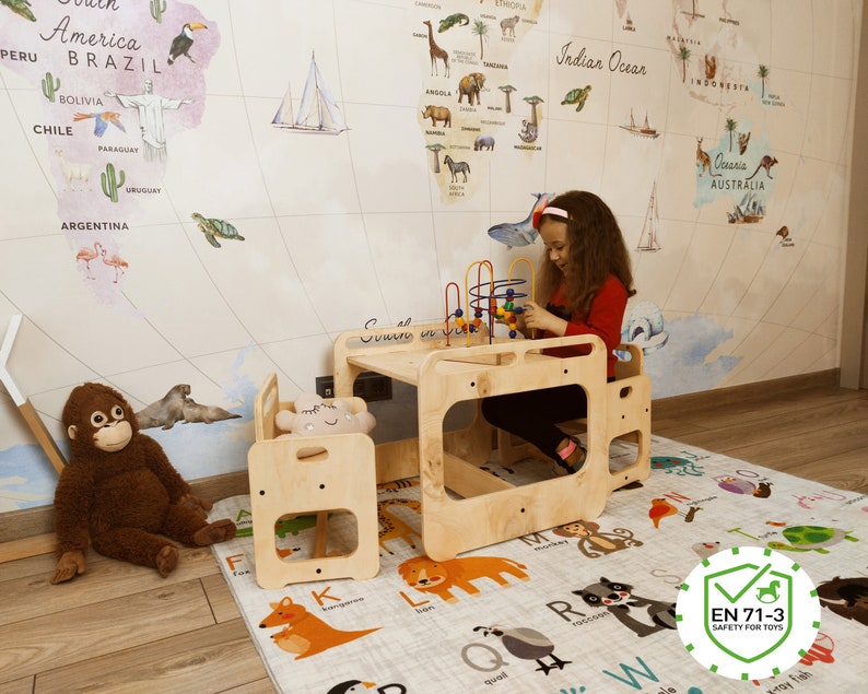 Puede incluir: Un conjunto de mesa y sillas de madera para jugar con un mapa del mundo en la pared detr&aacute;s. Un ni&ntilde;o peque&ntilde;o est&aacute; jugando con un juguete colorido. La mesa tiene un letrero que dice "Comer, dormir y jugar".