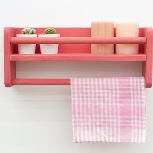 Può includere: Uno scaffale in legno rosa con due ripiani e un portasciugamani. Lo scaffale ha due piccole piante in vaso e due candele sul ripiano superiore. Un asciugamano da cucina a quadri rosa e bianco pende dal portasciugamani.