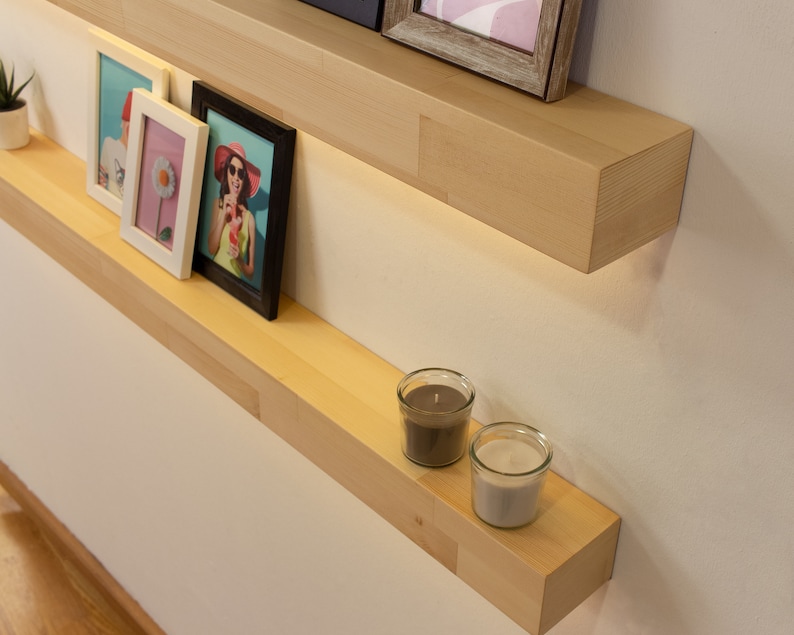 May include: Two light-colored wooden floating shelves mounted on a white wall. The top shelf holds framed pictures. The bottom shelf displays two glass candle holders and framed pictures. A small potted plant is on the top shelf.