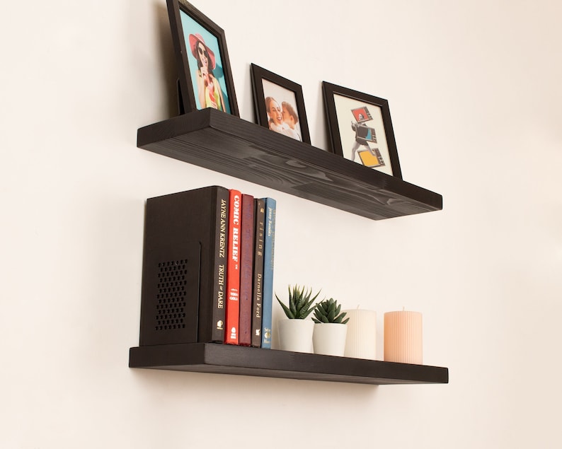 May include: Two black floating shelves with a black speaker, books, candles, and framed photos. The top shelf has three framed photos and the bottom shelf has a black speaker, six books, two small potted plants, and three candles.