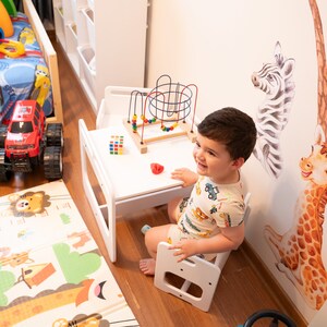May include: A white wooden table and chair set with a colorful toy bead maze and a red and white cube toy. A young child sits in the chair and smiles at the camera.