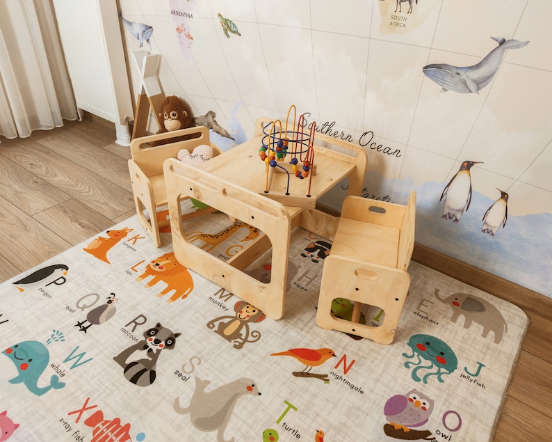 Puede incluir: Un conjunto de juegos de madera con dos sillas y una mesa. La mesa tiene un juguete colorido en la parte superior. El conjunto de juegos est&aacute; en una alfombra de juegos con un alfabeto e ilustraciones de animales.