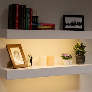 May include: Two white floating shelves with various decorative items. The top shelf holds books and a framed black and white cityscape. The bottom shelf displays a framed photo, candles, and small potted plants.