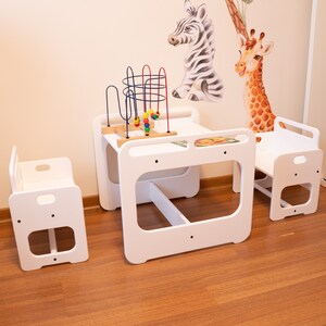 May include: White wooden children's table and chairs set with a colorful toy. The table has a built-in shelf and the chairs have a simple design.