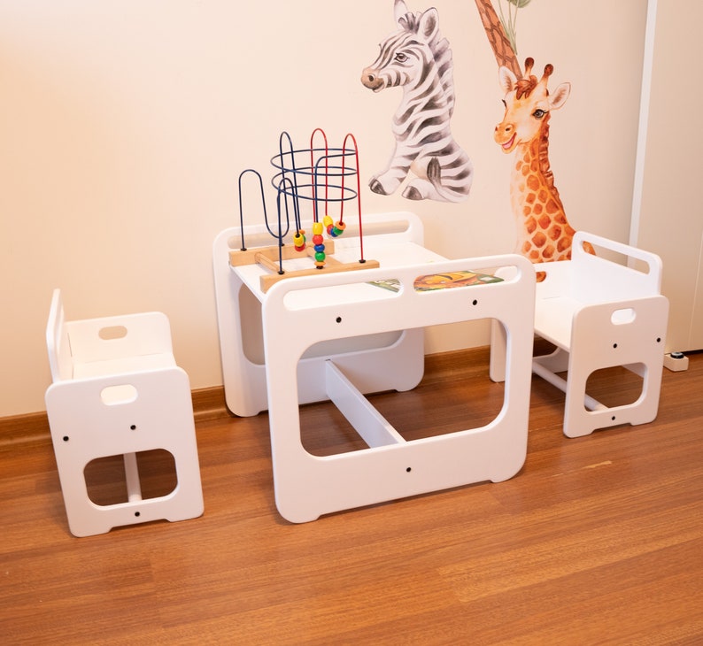 May include: White wooden children's table and chairs set with a colorful toy. The table has a built-in shelf and the chairs have a simple design.