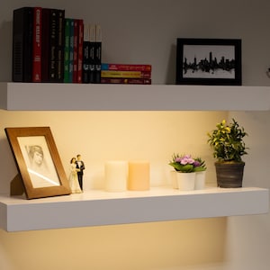 May include: Two white floating shelves with various decorative items. The top shelf holds books and a framed skyline print. The bottom shelf displays a framed photo, candles, and potted plants. The shelves are illuminated from below.
