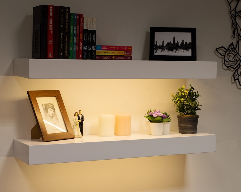 May include: Two white floating shelves with various decorative items. The top shelf holds books and a framed skyline print. The bottom shelf displays a framed photo, candles, and potted plants. The shelves are illuminated from below.