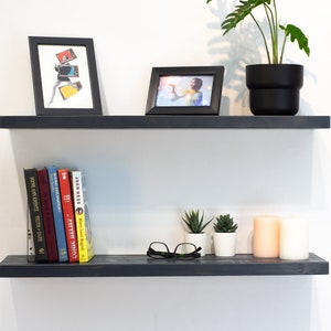 Könnte beinhalten: Zwei schwarze schwebende Regale mit verschiedenen Gegenständen darauf, darunter Bücher, Pflanzen, Kerzen und gerahmte Fotos. Die Regale sind an einer weißen Wand montiert.