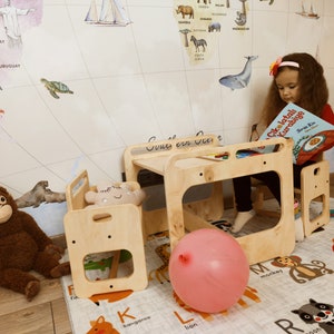 Puede incluir: Un juego de mesa y sillas de madera clara para ni&ntilde;os con un globo rosa en el suelo. La mesa tiene un peque&ntilde;o estante debajo y las sillas tienen un respaldo peque&ntilde;o y un recorte para los pies de un ni&ntilde;o. Un ni&ntilde;o est&aacute; sentado en la mesa leyendo un libro titulado "&Ccedil;ikolatalı Kurabiye".