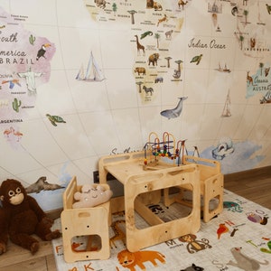 Puede incluir: Un conjunto de mesa y sillas de madera para jugar con un papel tapiz de mapa del mundo en el fondo. La mesa tiene un estante incorporado y las sillas tienen un peque&ntilde;o coj&iacute;n. El conjunto de juego est&aacute; sobre una alfombra de alfabeto blanca y gris.