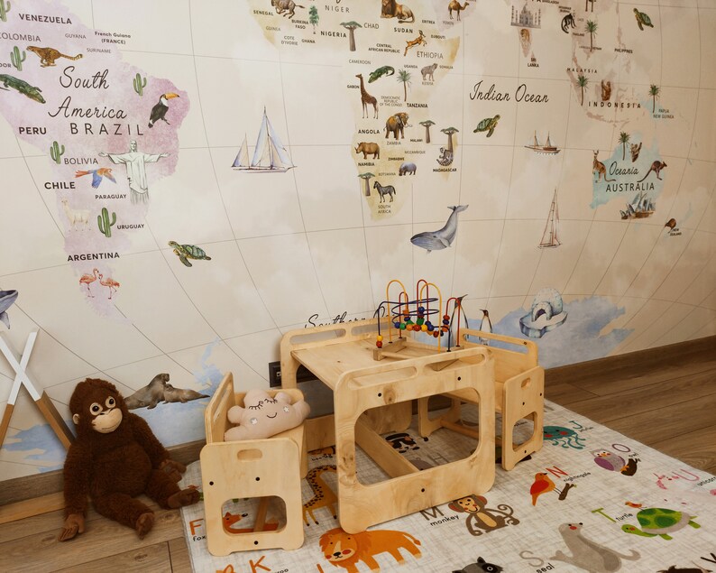 Puede incluir: Un conjunto de mesa y sillas de madera para jugar con un papel tapiz de mapa del mundo en el fondo. La mesa tiene un estante incorporado y las sillas tienen un peque&ntilde;o coj&iacute;n. El conjunto de juego est&aacute; sobre una alfombra de alfabeto blanca y gris.