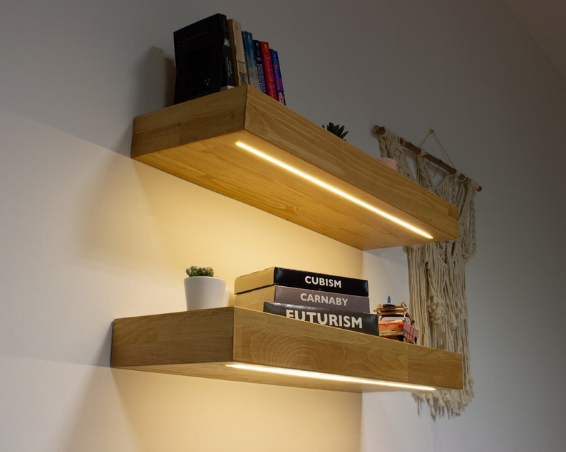 May include: Two wooden floating shelves with integrated LED lighting. The shelves are made of light-colored wood and are mounted on a white wall. Books, a small potted plant, and decorative items are displayed on the shelves.