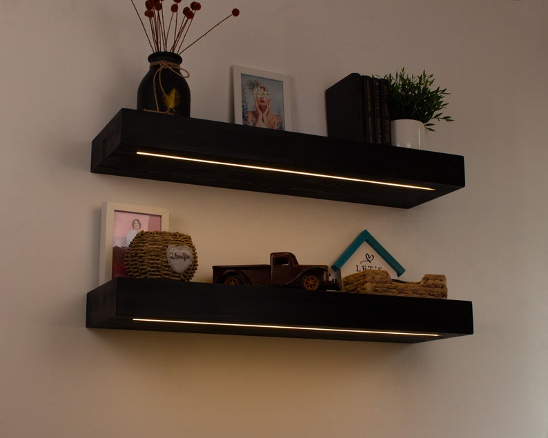 K&ouml;nnte beinhalten: Zwei schwarze schwebende Regale mit integrierter LED-Beleuchtung an einer wei&szlig;en Wand. Die Regale zeigen dekorative Gegenst&auml;nde wie eine Vase mit roten Beeren, gerahmte Fotos, einen kleinen LKW und eine Topfpflanze. Die Regale sind rechteckig.
