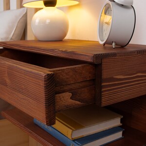 May include: A dark brown wooden floating nightstand featuring a single horizontal drawer with a visible wood grain pattern. The nightstand has an open storage shelf underneath, currently holding two stacked books with blue and yellow covers.