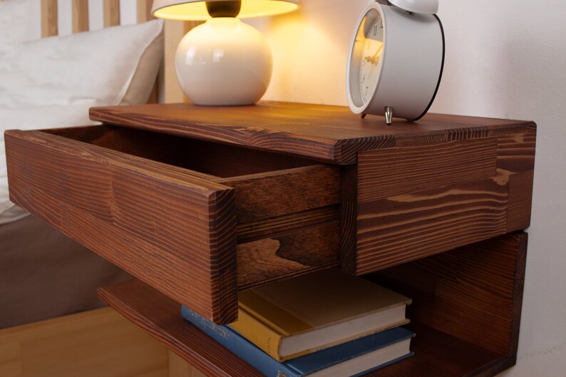 May include: A dark brown wooden floating nightstand featuring a single horizontal drawer with a visible wood grain pattern. The nightstand has an open storage shelf underneath, currently holding two stacked books with blue and yellow covers.