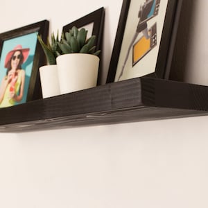 May include: Black floating shelf with three framed pictures and two potted plants. The shelf is mounted on a white wall.