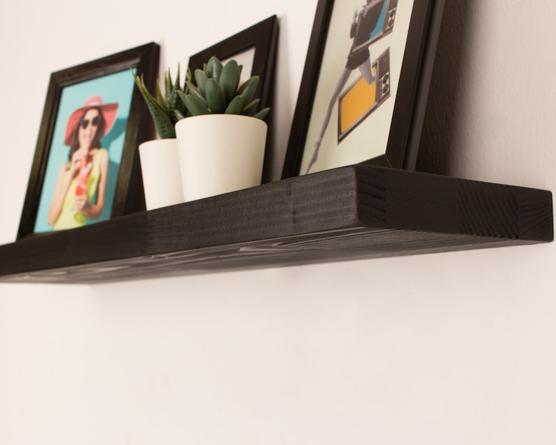 May include: Black floating shelf with three framed pictures and two potted plants. The shelf is mounted on a white wall.