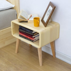 May include: A light wood nightstand with rounded edges and four legs. The nightstand has a shelf for storing books and a small picture frame on top. A yellow mug and a book are also on top of the nightstand.