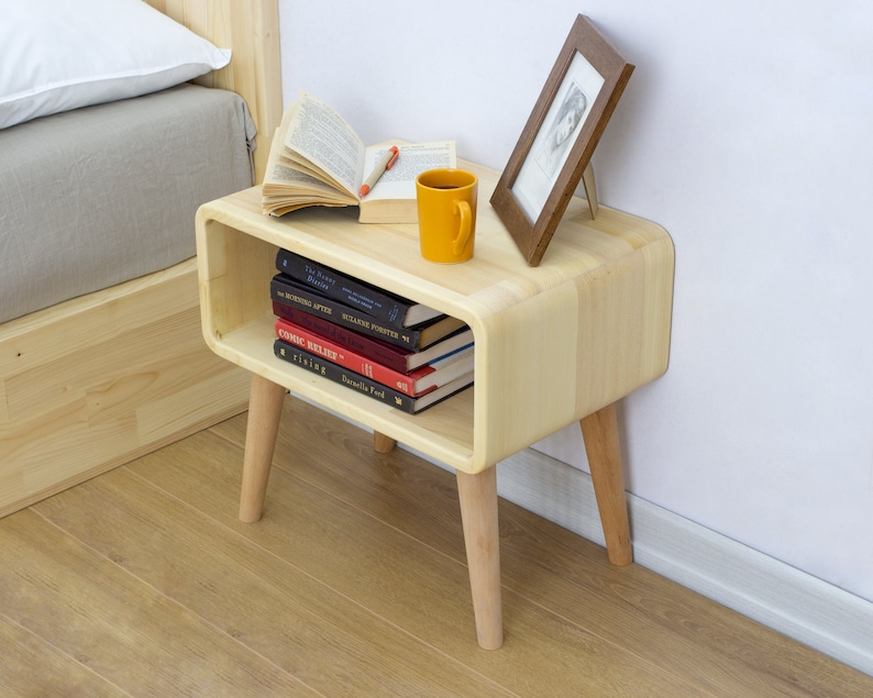 May include: A light wood nightstand with rounded edges and four legs. The nightstand has a shelf for storing books and a small picture frame on top. A yellow mug and a book are also on top of the nightstand.