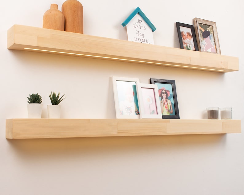 May include: Two light-colored wooden floating shelves mounted on a white wall. The top shelf has a built-in light and displays wooden vases, a small house-shaped sign that says "Let's stay home", and framed photos. The bottom shelf holds small potted plants and more framed photos.