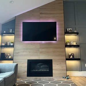 K&ouml;nnte beinhalten: Ein modernes Wohnzimmer mit Kamin und gro&szlig;em Flachbildfernseher. Der Fernseher ist an einer Akzentwand aus Holz mit einem rosa LED-Lichtrand befestigt. Schwarze Regale mit Dekoration befinden sich auf beiden Seiten des Fernsehers. Im Vordergrund befindet sich ein graues Sofa.