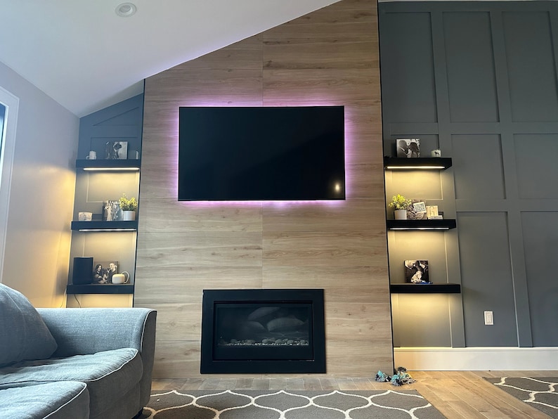 K&ouml;nnte beinhalten: Ein modernes Wohnzimmer mit Kamin und gro&szlig;em Flachbildfernseher. Der Fernseher ist an einer Akzentwand aus Holz mit einem rosa LED-Lichtrand befestigt. Schwarze Regale mit Dekoration befinden sich auf beiden Seiten des Fernsehers. Im Vordergrund befindet sich ein graues Sofa.