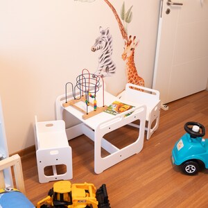 May include: A white wooden children's table and chairs set with a colorful toy and a book. The set is on a wooden floor with a blue toy car and a yellow toy truck.
