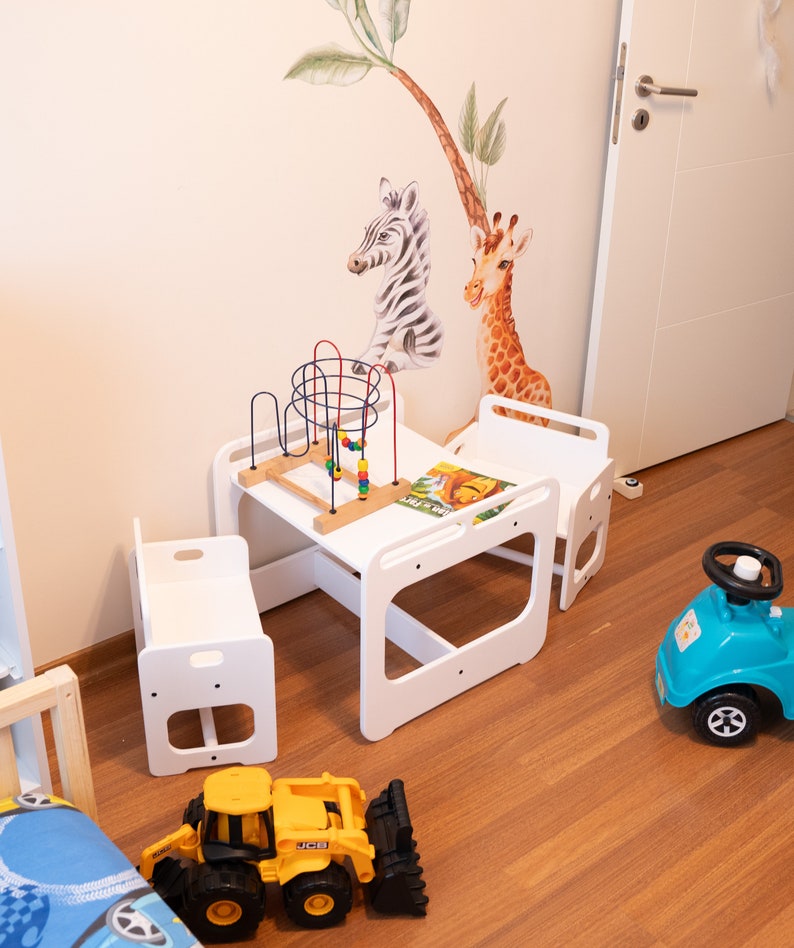 May include: A white wooden children's table and chairs set with a colorful toy and a book. The set is on a wooden floor with a blue toy car and a yellow toy truck.