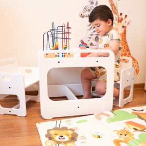May include: A white wooden children's table and chairs set with a colorful bead maze toy. The table has a small shelf underneath. The chairs have a step up to help little ones get in and out.