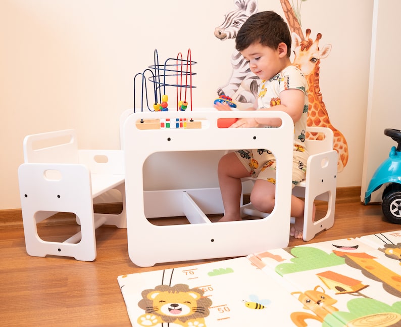 May include: A white wooden children's table and chairs set with a colorful bead maze toy. The table has a small shelf underneath. The chairs have a step up to help little ones get in and out.