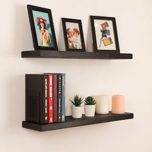 May include: Two black floating shelves with a variety of items on them. The top shelf has three framed pictures, one with a woman in a red hat, one with a couple, and one with a person holding a stack of records. The bottom shelf has a black speaker, a row of books, and three candles.