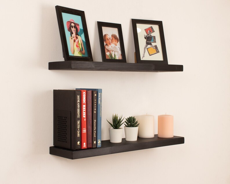 May include: Two black floating shelves with a variety of items on them. The top shelf has three framed pictures, one with a woman in a red hat, one with a couple, and one with a person holding a stack of records. The bottom shelf has a black speaker, a row of books, and three candles.